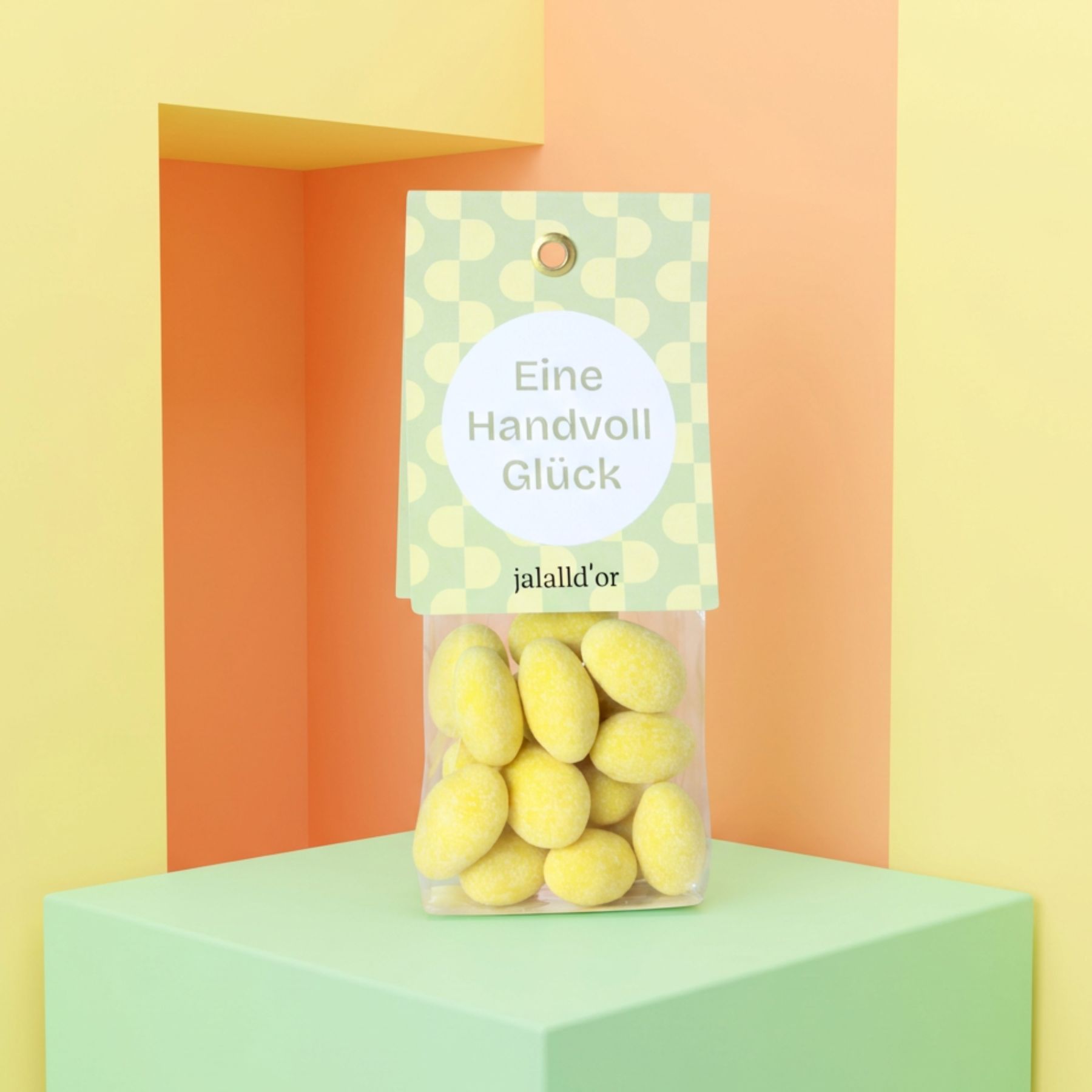 Zitronen-Schoko-Mandeln in Geschenktüte auf hellgrünem Podest mit gelb-orangem Hintergrund