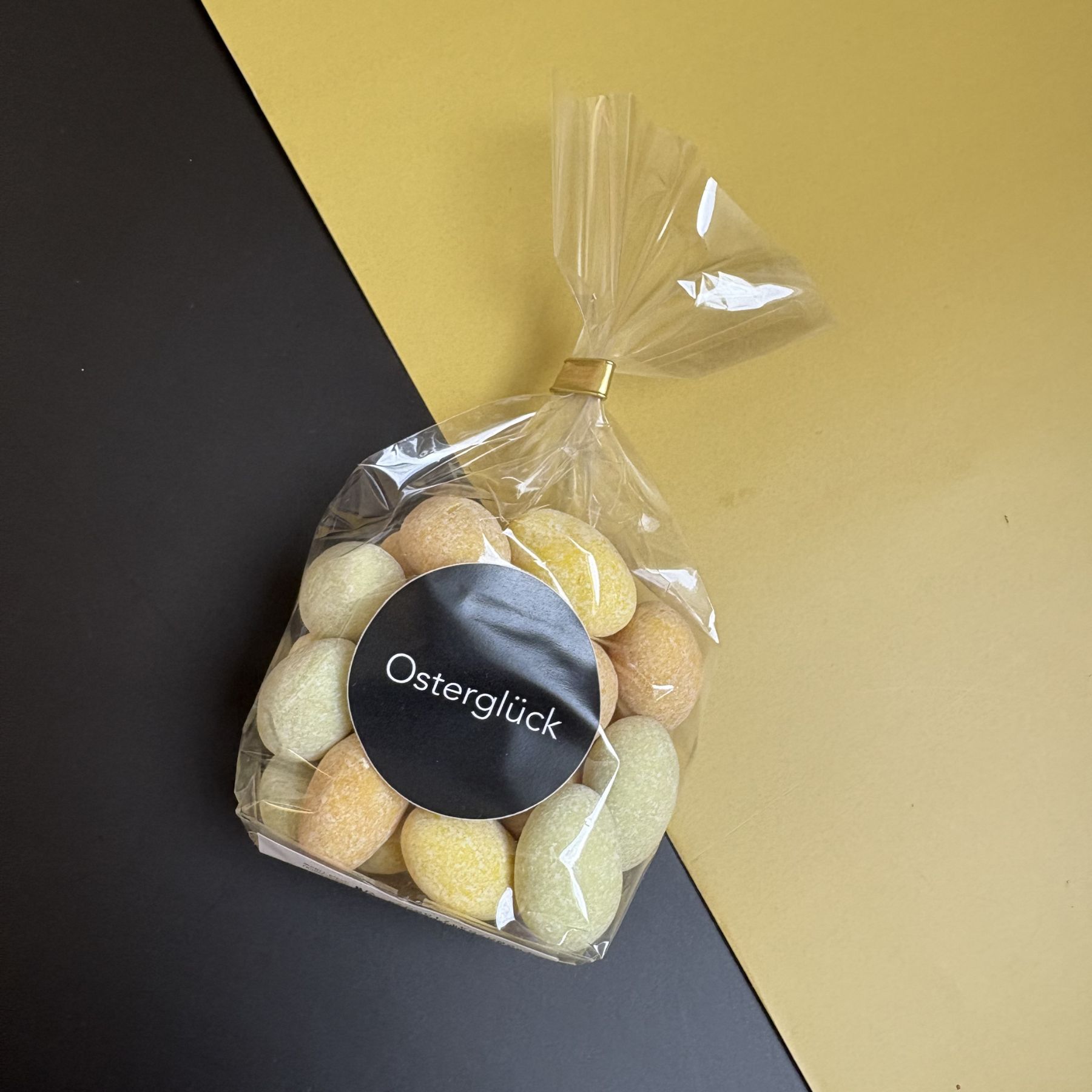 Cellotüte mit Aufkleber Osterglück und gelb-orange-hellgrünen Schokomandeln liegt auf diagonal zweifarbigem Untergrund in schwarz-gelb